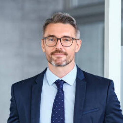 Portrait of handsome mid adult business man standing in modern office. Successful mature entrepreneur in formal clothing looking at camera with satisfaction. Confident man in suit with eyeglasses and beard standing with hands in pocket and looking at camera in office.